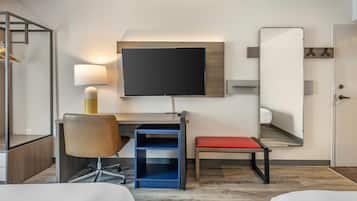 Una habitación de hotel con una cama, un escritorio con silla, un televisor, un espejo y un banco rojo.