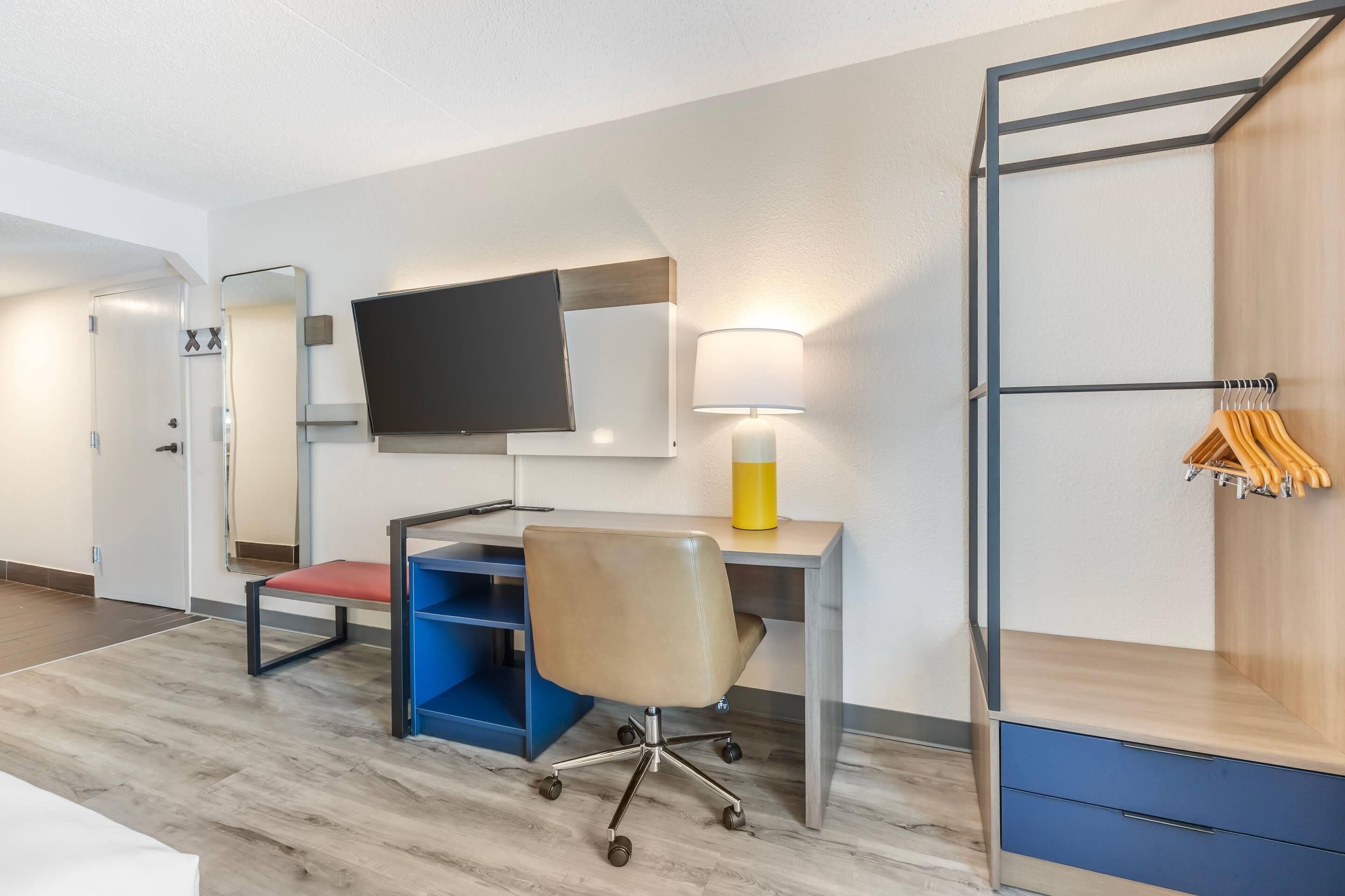 A hotel room with a desk, chair, TV, lamp, and a wardrobe with hanging clothes.
