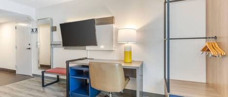 A hotel room with a desk, chair, TV, lamp, and a wardrobe with hanging clothes.