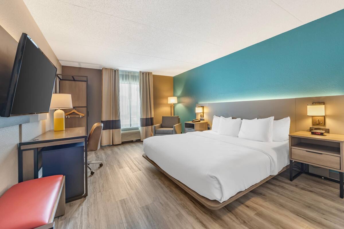 A hotel room with a large bed, a desk with a chair, a TV, and a desk lamp.
