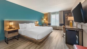 Una habitación de hotel con cama, mesitas de noche, escritorio, silla, televisor y ventana con cortinas.