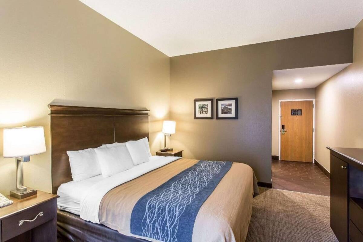 A hotel room with a bed, bedside lamps, a desk with a lamp, a dresser, and a door leading to another room.