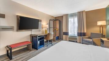 A hotel room with a large bed, a desk with a chair, a TV mounted on the wall, a bedside table with a lamp, and a window with curtains.