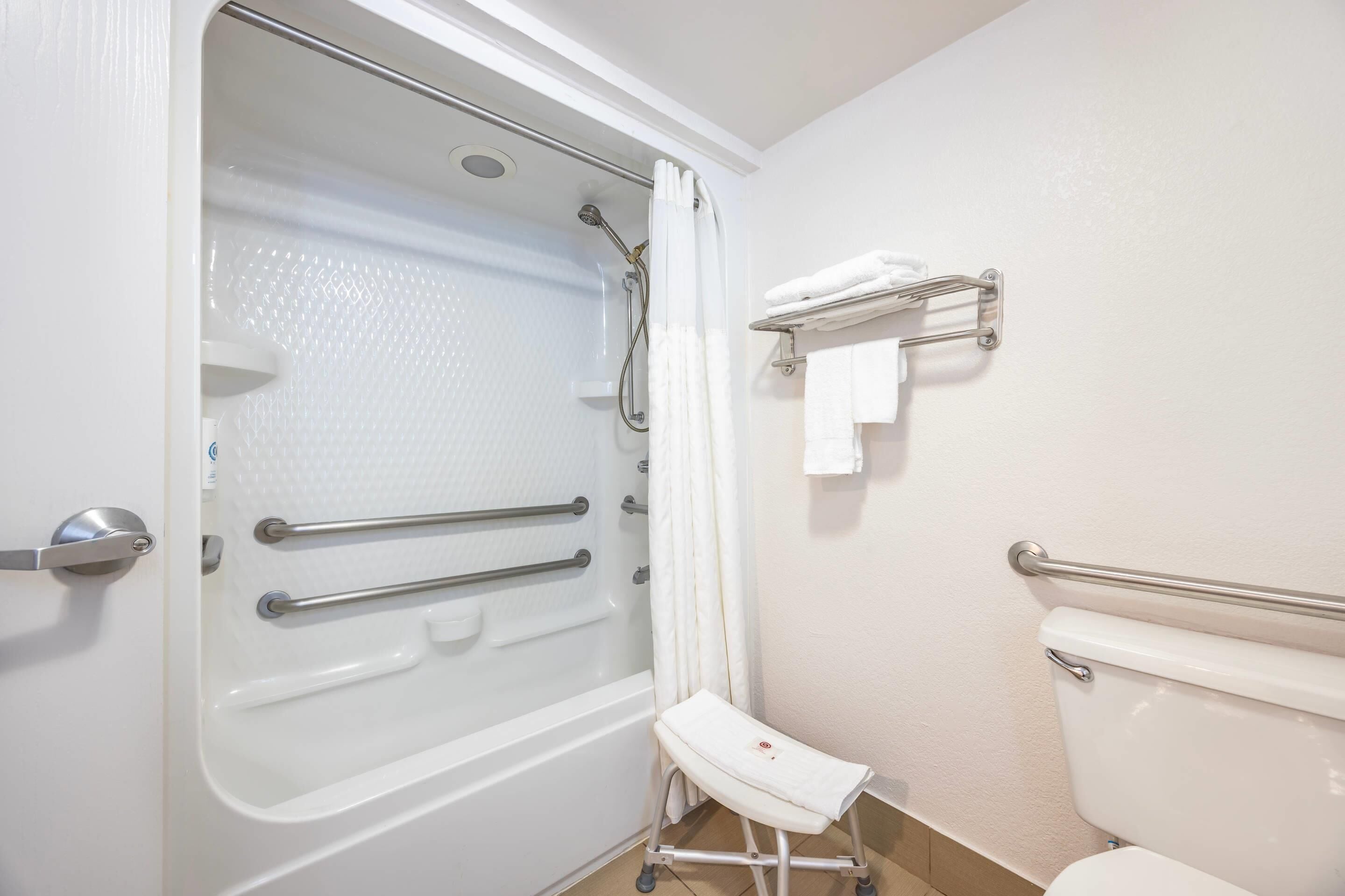 Un baño con bañera, ducha e inodoro. Incluye barras de apoyo y un asiento para la ducha.