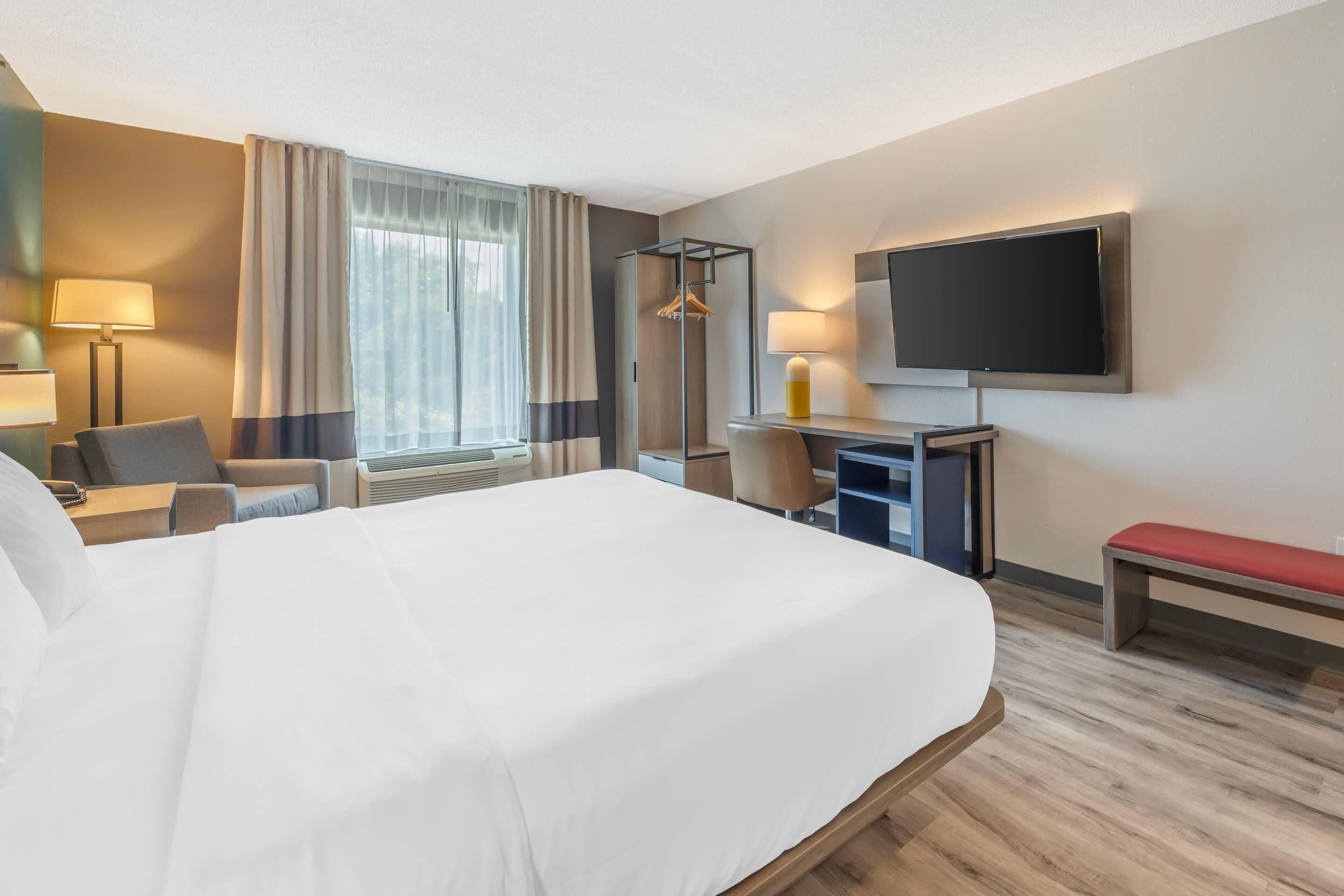 Habitación de hotel con una cama grande, un escritorio con silla, un televisor montado en la pared y una ventana con cortinas.