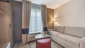 Habitación de hotel con sofá, una mesita, una maleta roja, una lámpara y una ventana con cortinas.