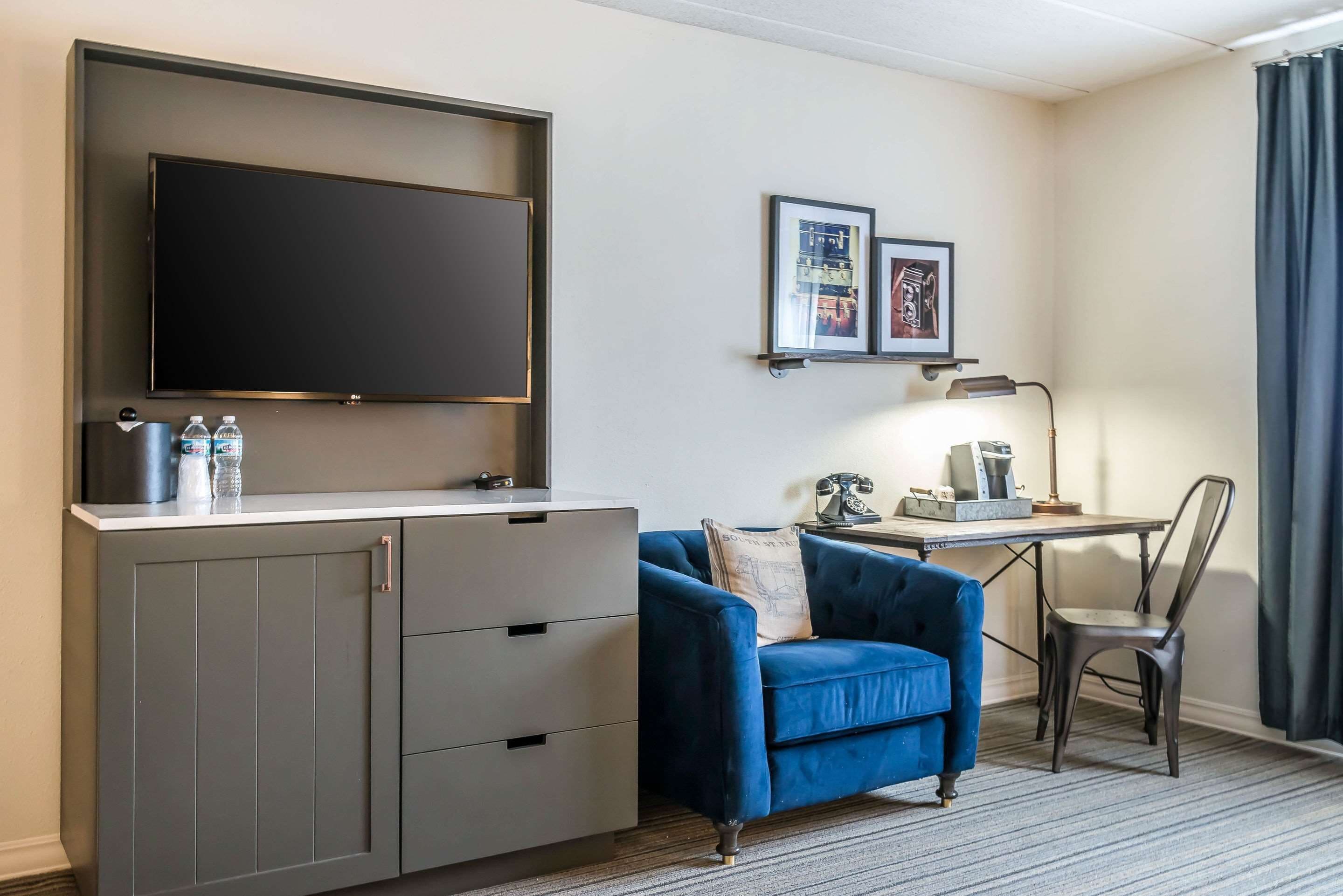 Quarto de hotel com uma televisão de tela grande fixada na parede, uma poltrona azul, uma escrivaninha com abajur e uma cadeira.