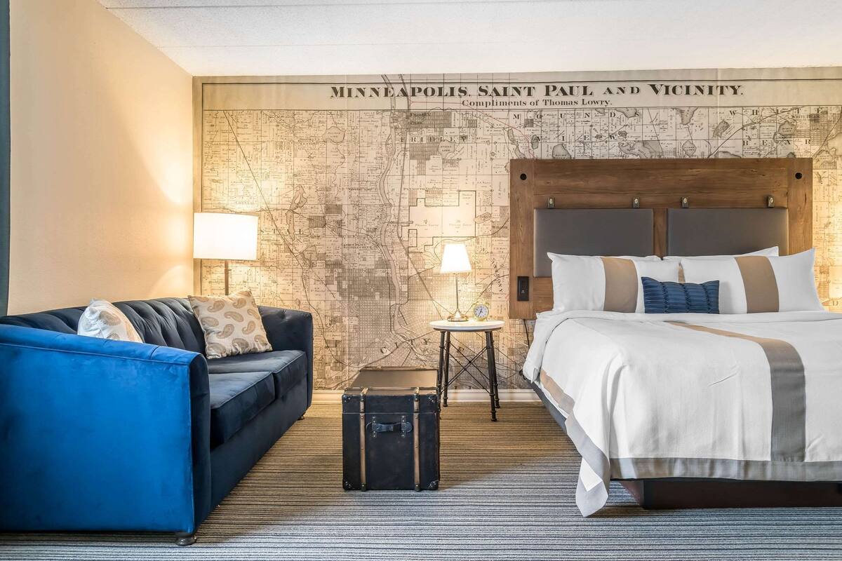 Ein Hotelzimmer mit einer großen Karte an der Wand, einem Bett, einem blauen Sessel, einem kleinen Tisch mit einer Lampe und einem Koffer.