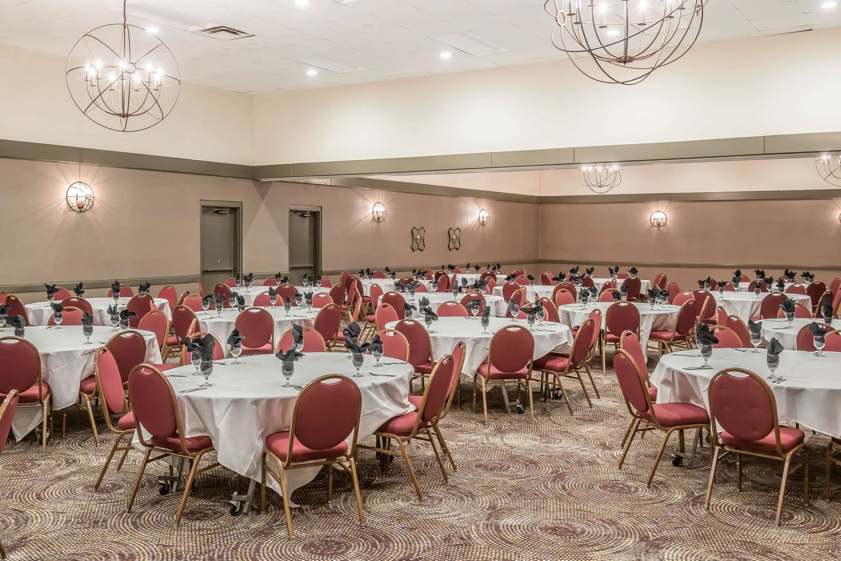 Un amplio salón de banquetes con varias mesas redondas, cada una con sillas rojas y manteles blancos, y candelabros colgando del techo.