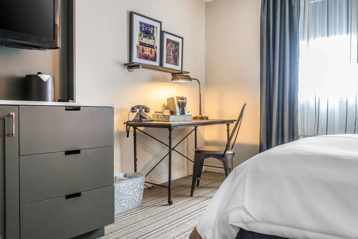 Ein Hotelzimmer mit einem Bett, einem Schreibtisch mit Lampe, einem Sessel, einer Minibar, einem Mülleimer und einem an der Wand befestigten Fernseher.