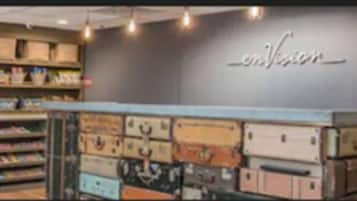 A store interior with shelves stocked with various items, a counter with suitcases, and a wall with the word 'envision' displayed.