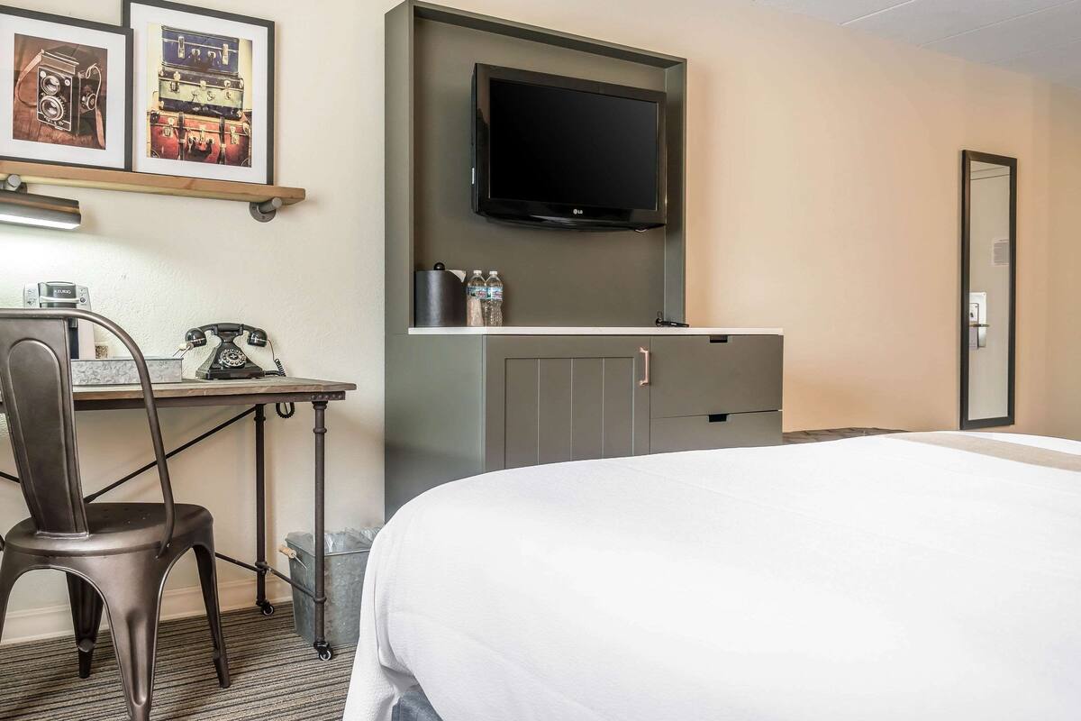 Ein Hotelzimmer mit einem Bett, einem Schreibtisch mit Stuhl, einem Telefon, einem an der Wand befestigten Fernseher und einem Papierkorb.