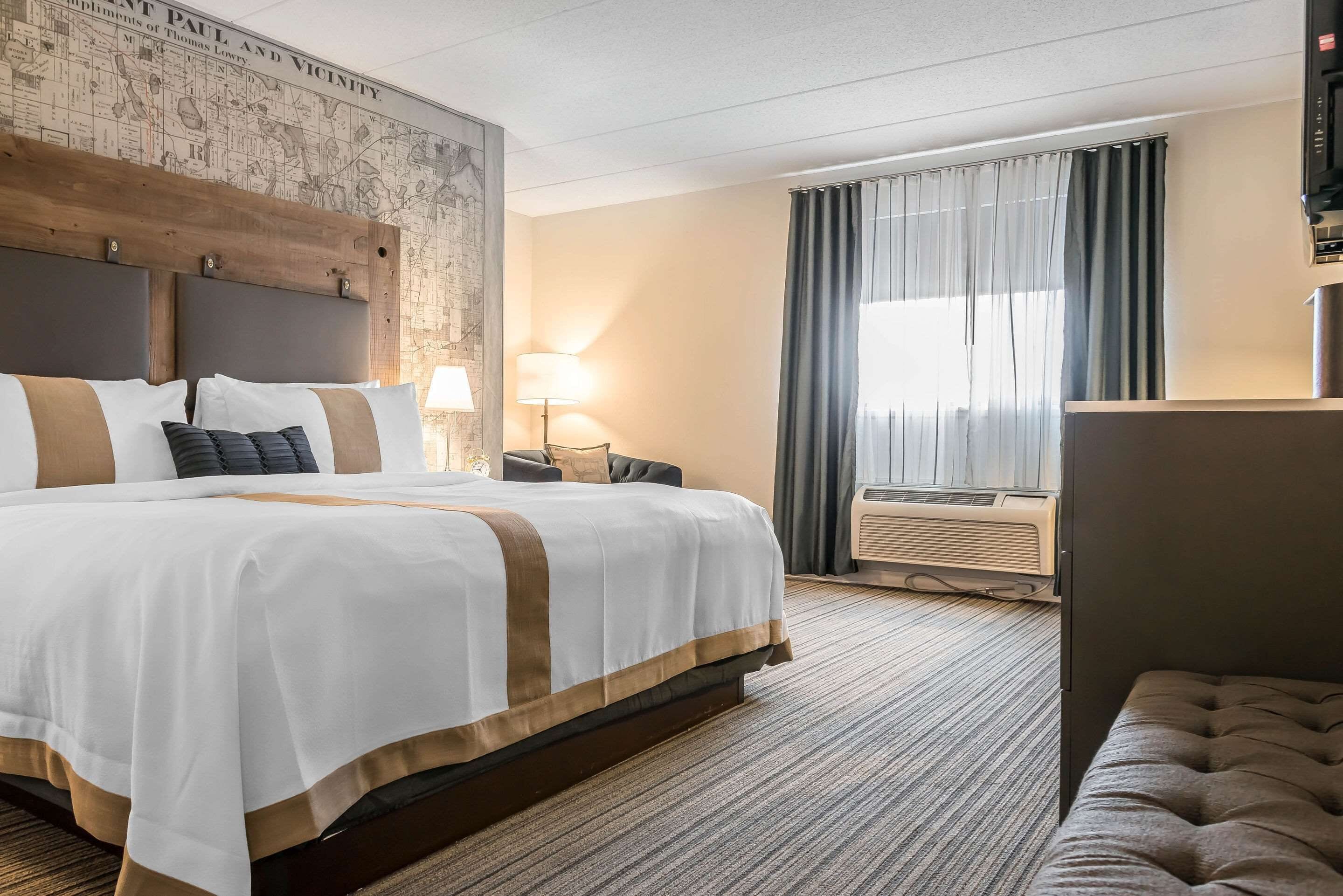 A hotel room with a large bed, a headboard with a map, an air conditioning unit, and a window with curtains.