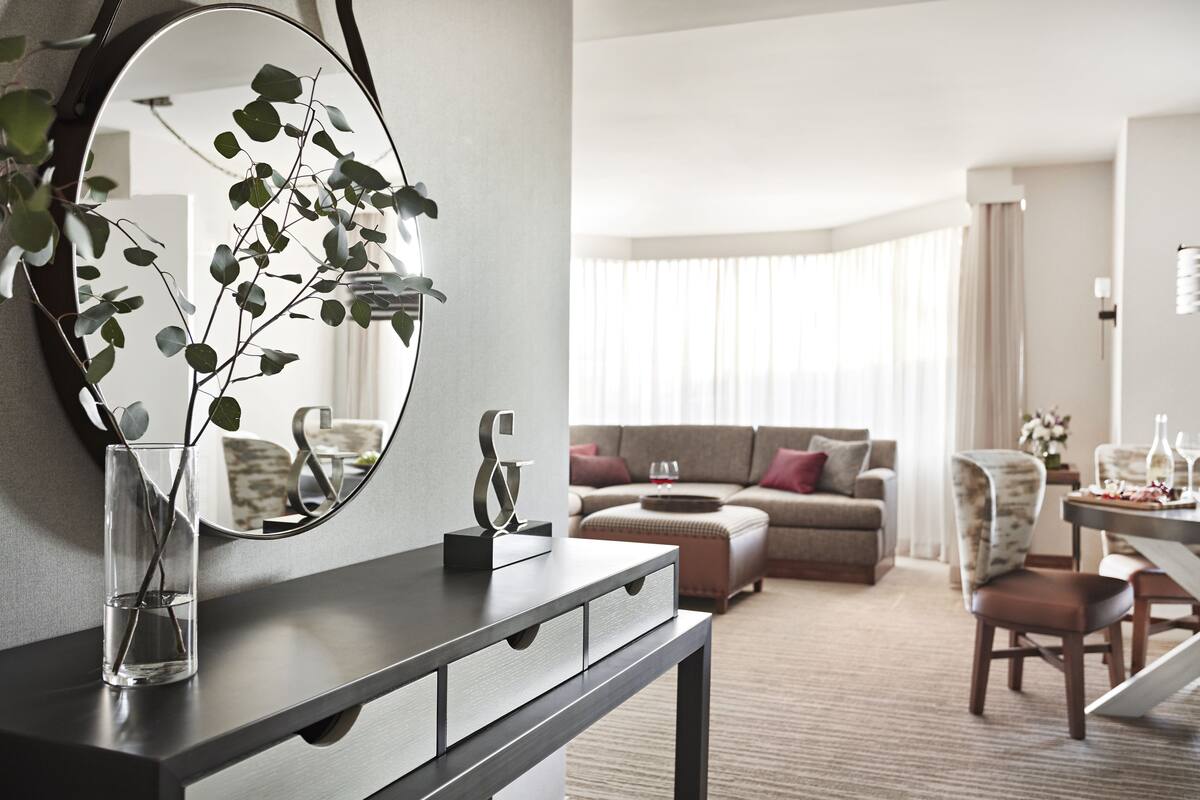 A modern hotel room with a dark wood vanity, a large mirror, and a glass vase with green leaves. There is a beige sofa, a dining table with chairs, and a window with sheer curtains.