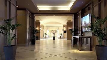 Un hall de hotel con piso de baldosas, plantas grandes en macetas, un mostrador de recepción y una pantalla de televisión.