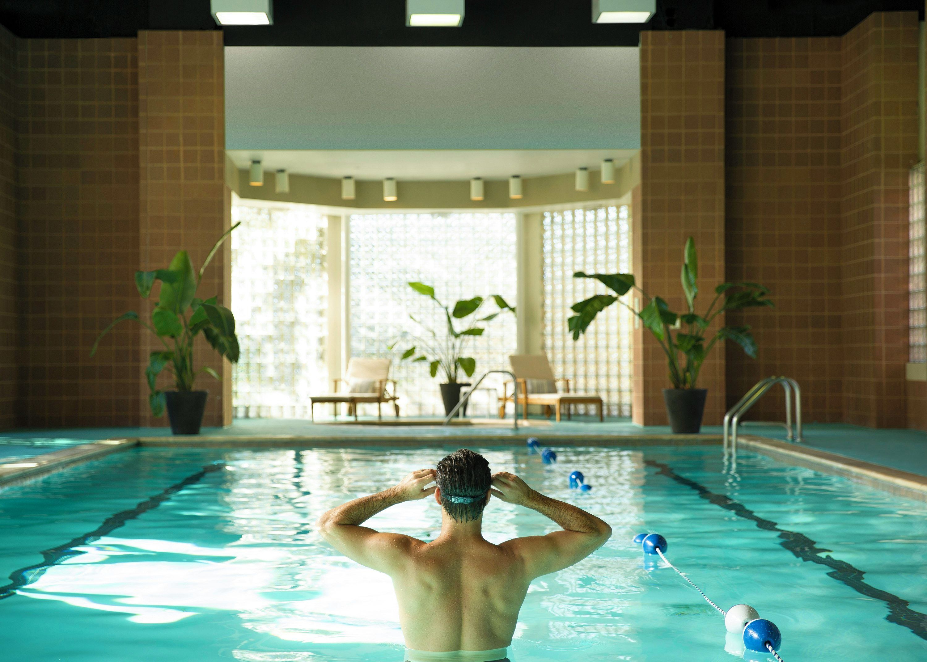 An indoor swimming pool with a person in the water, surrounded by potted plants and chairs.