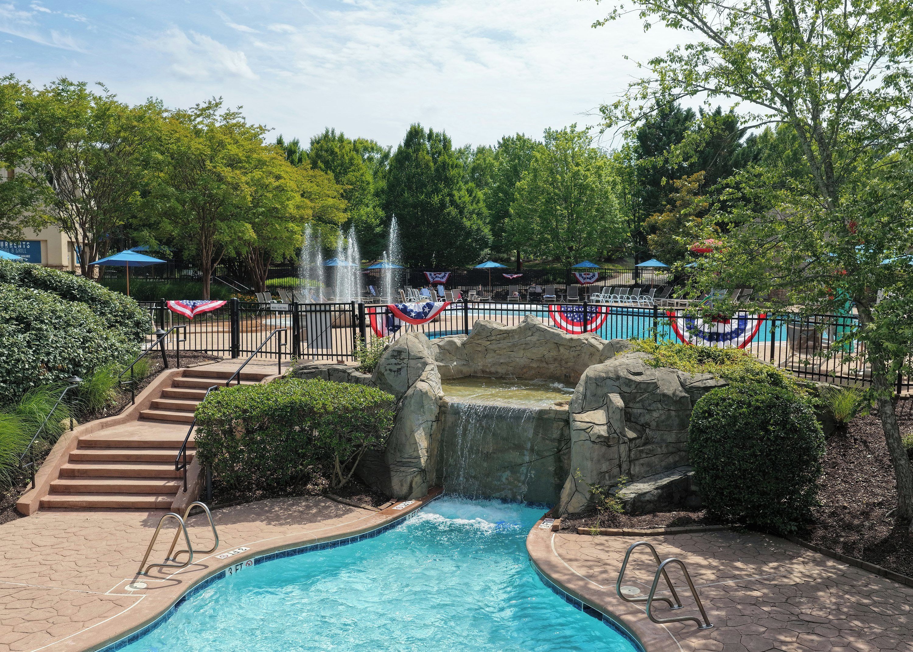A swimming pool with a waterfall feature, surrounded by a fence with American flag decorations, and a set of stairs leading up to the pool area.