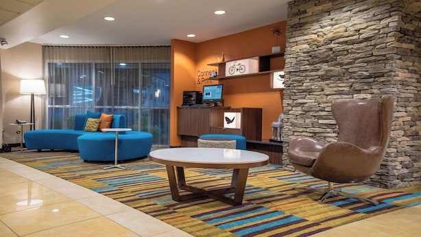 A modern hotel lobby with a blue sofa, a round table, a stone accent wall, and a reception desk with a computer and a bicycle sign. - Fairfield Inn & Suites by Marriott Knoxville/East (Knoxville)
