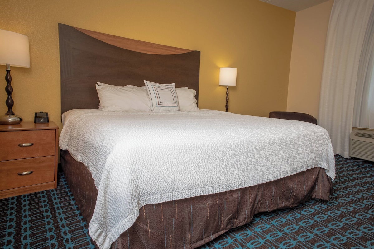 A hotel room with a large bed, a wooden nightstand with a lamp, a chair, and a wall-mounted heater.