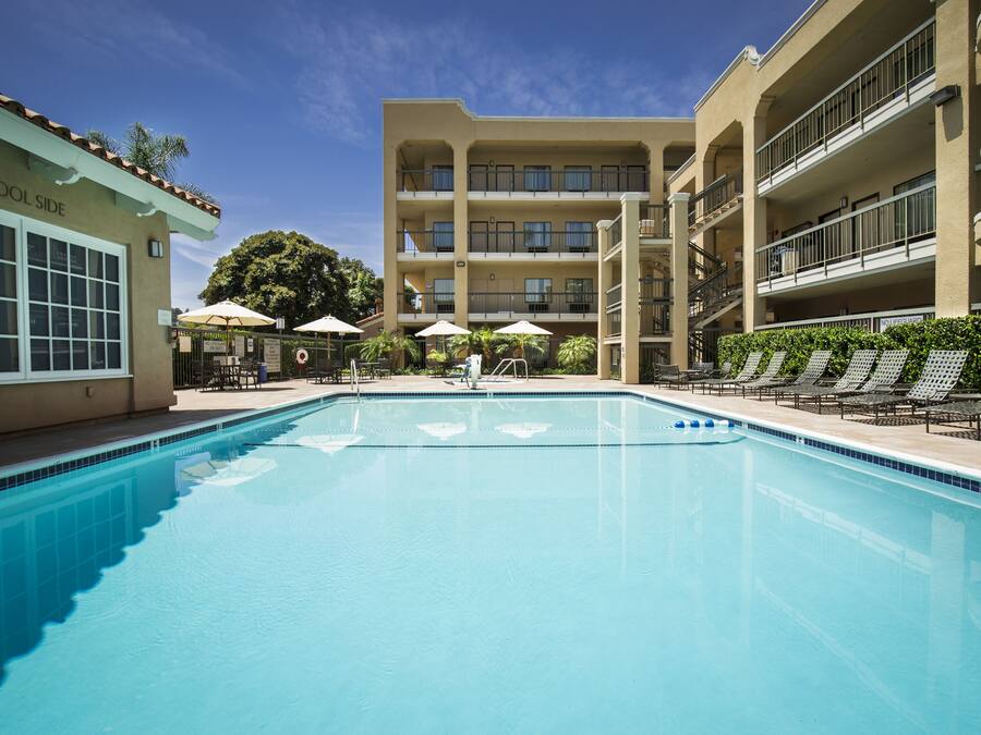 Una piscina con sillones de descanso y sombrillas, rodeada por un edificio de varios pisos con balcones.
