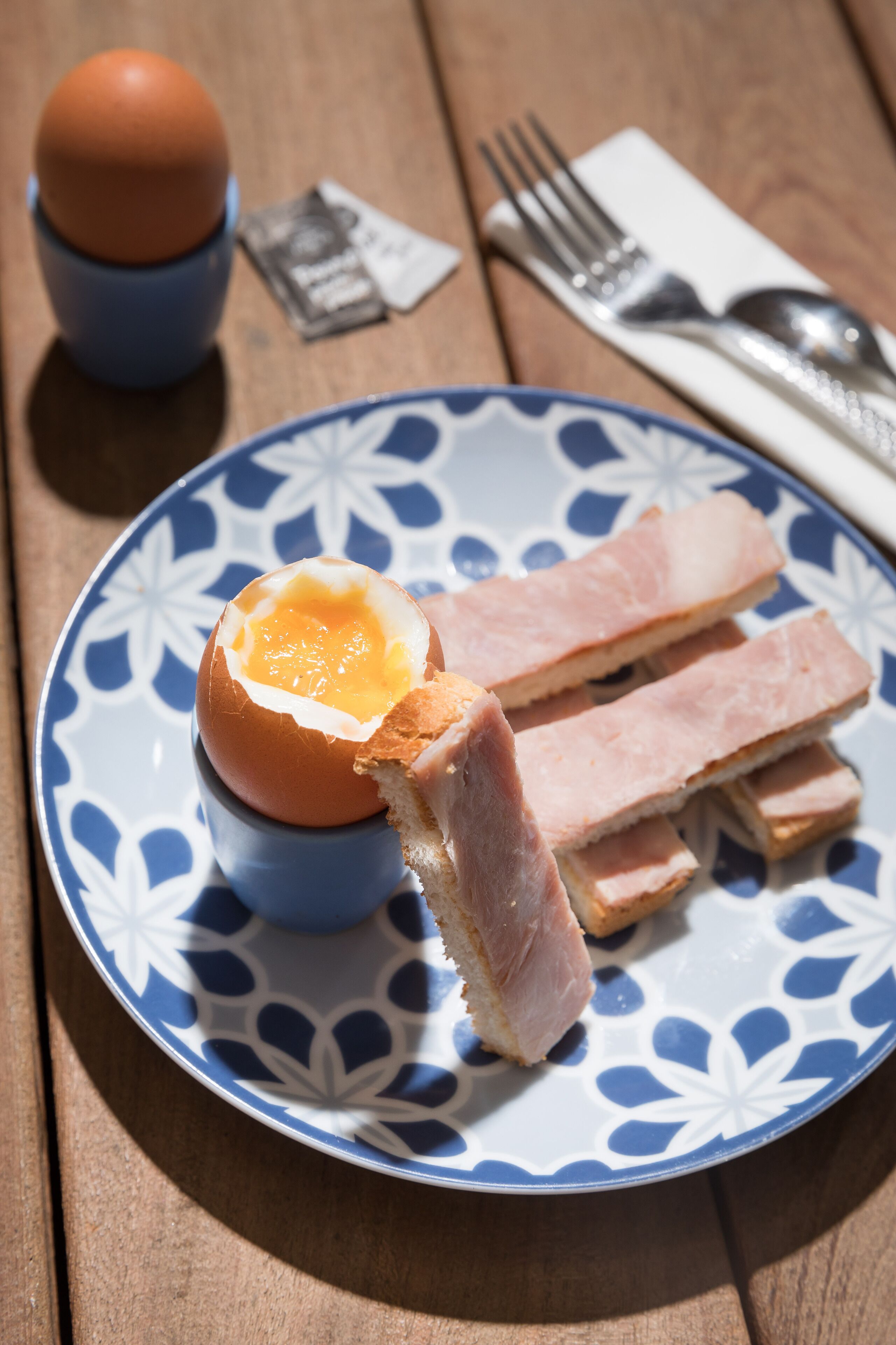A plate with a boiled egg, ham slices, and a blue and white patterned plate on a wooden table.