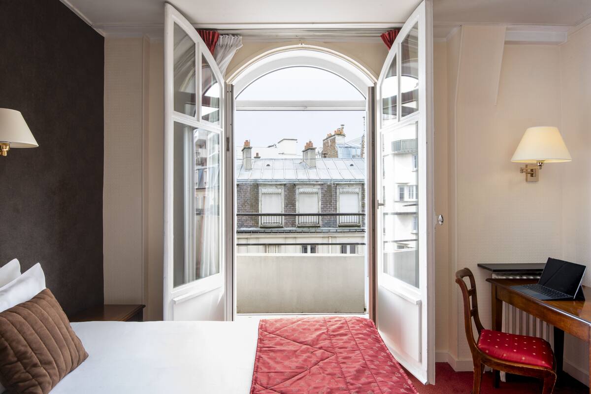 Ein Hotelzimmer mit Bett, Schreibtisch mit Laptop und offener Tür zu einem Balkon mit Blick auf Gebäude.