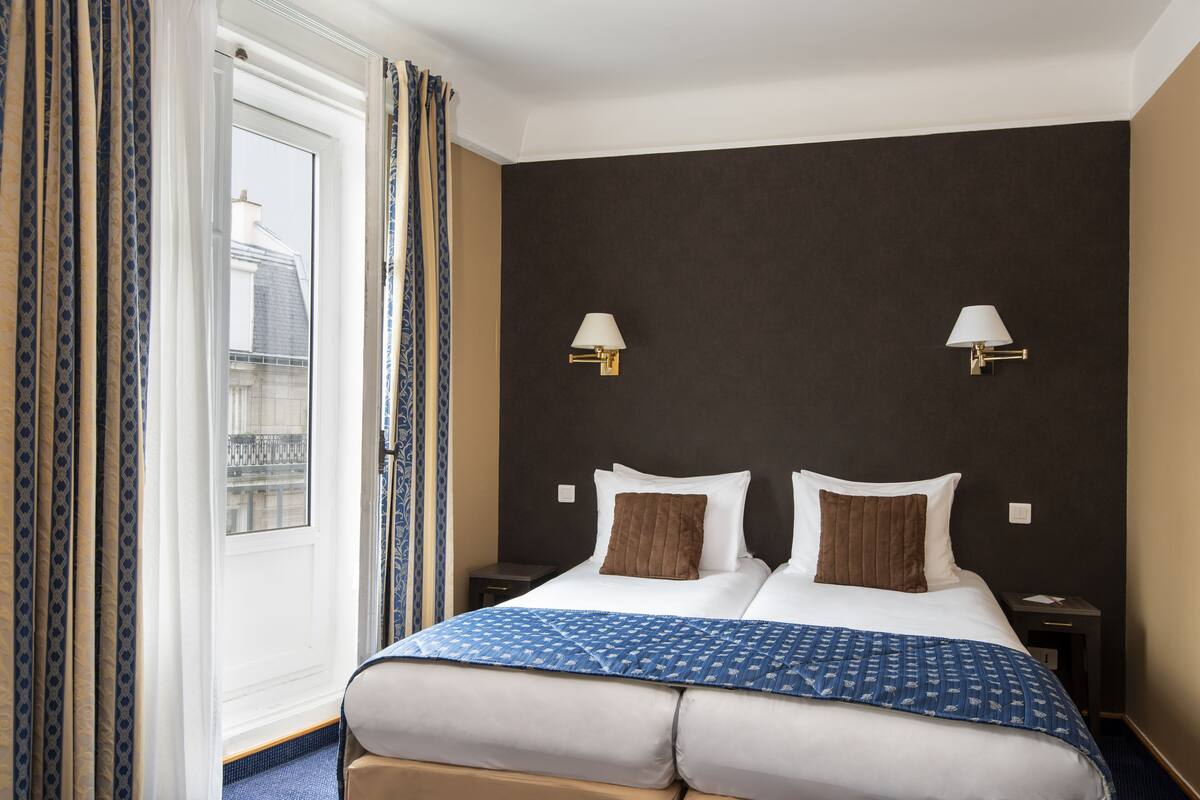 Ein Hotelzimmer mit einem großen Bett, zwei Nachttischen, einem Fenster mit Vorhängen und zwei Wandlampen.