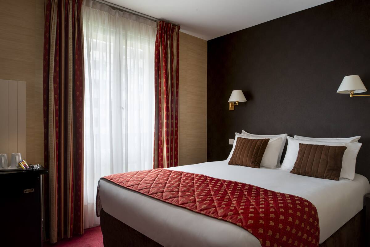 Ein Hotelzimmer mit einem großen Bett, zwei Nachttischlampen, einem Schreibtisch mit Telefon und einem Fenster mit durchsichtigen Vorhängen.