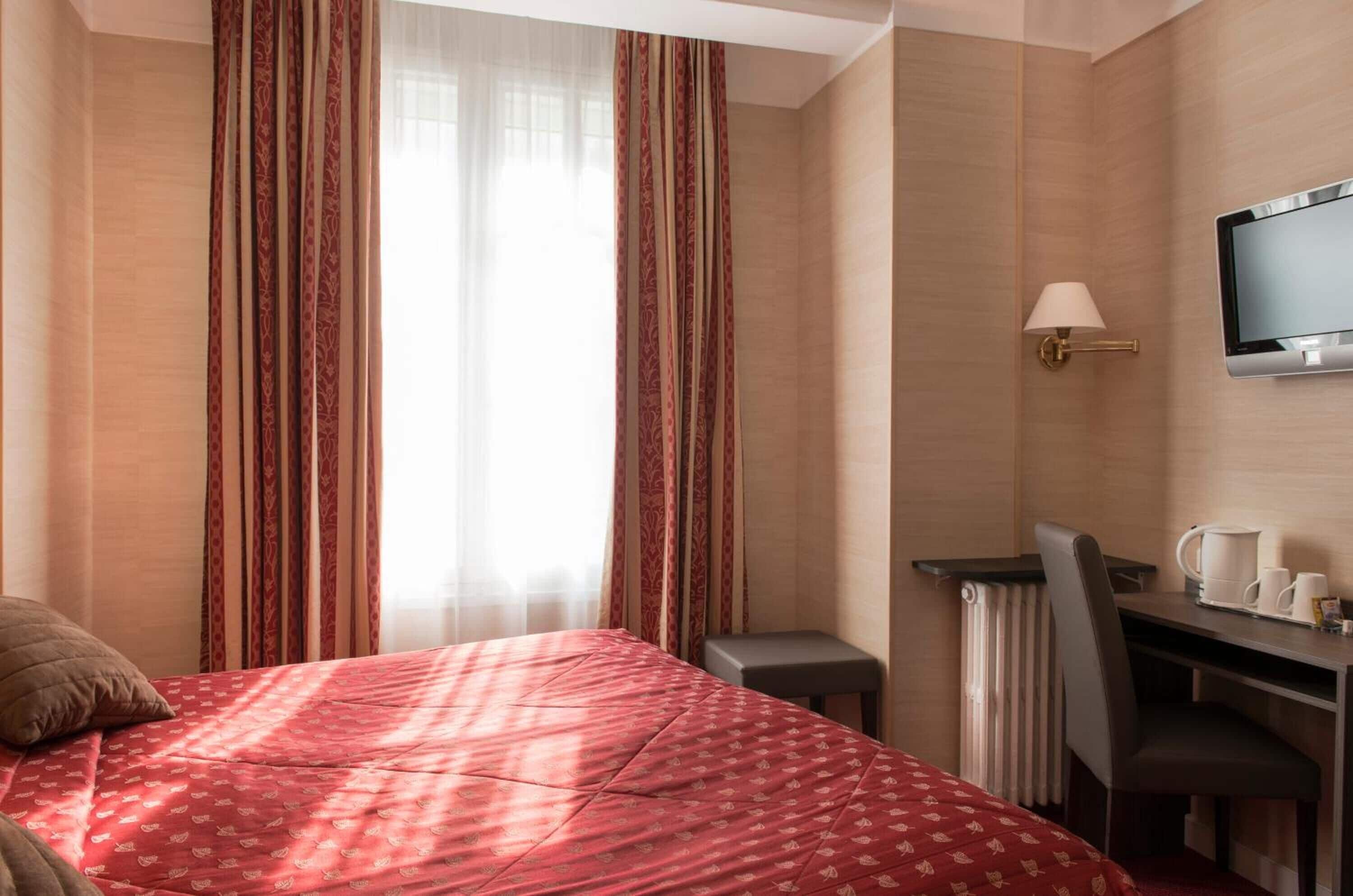 Une chambre d’hôtel avec un lit, un bureau, une chaise, une télévision et une fenêtre avec des rideaux.