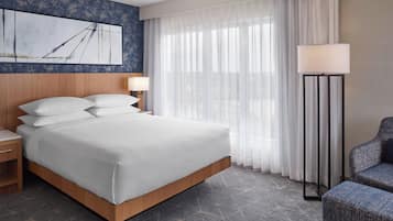 Habitación de hotel con cama, mesita de noche, lámpara, silla y un ventanal con cortinas.