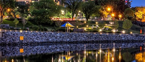 Un immeuble de grande hauteur, doté de nombreux étages, un jardin bien entretenu avec des arbres et des arbustes, et une étendue d’eau calme qui reflète l’immeuble et les lumières.