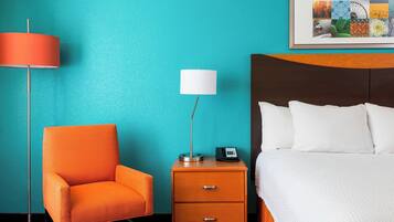 A hotel room with a teal wall, a bed with white bedding, an orange armchair, a nightstand with a lamp, and a framed picture above the bed.