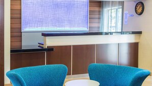 Ett välkomnande område med modern design, med en receptionsdisk, en digital skärm och två blå fåtöljer placerade runt ett litet runt bord.