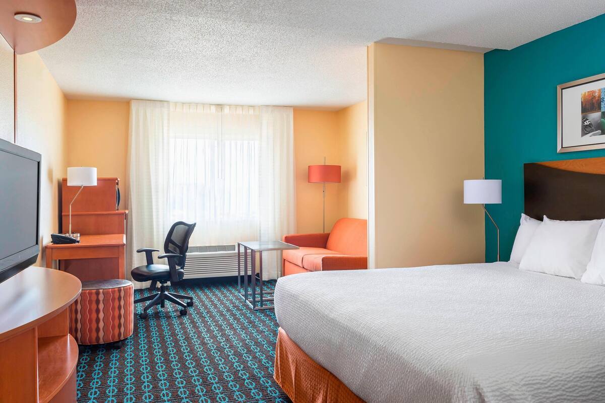 A hotel room with a bed, a desk, a chair, a small table, and a lamp.