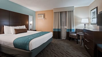 Ein Hotelzimmer mit einem großen Bett, einem Schreibtisch, einem Sessel, einem Fernseher und einem Fenster mit Vorhängen.