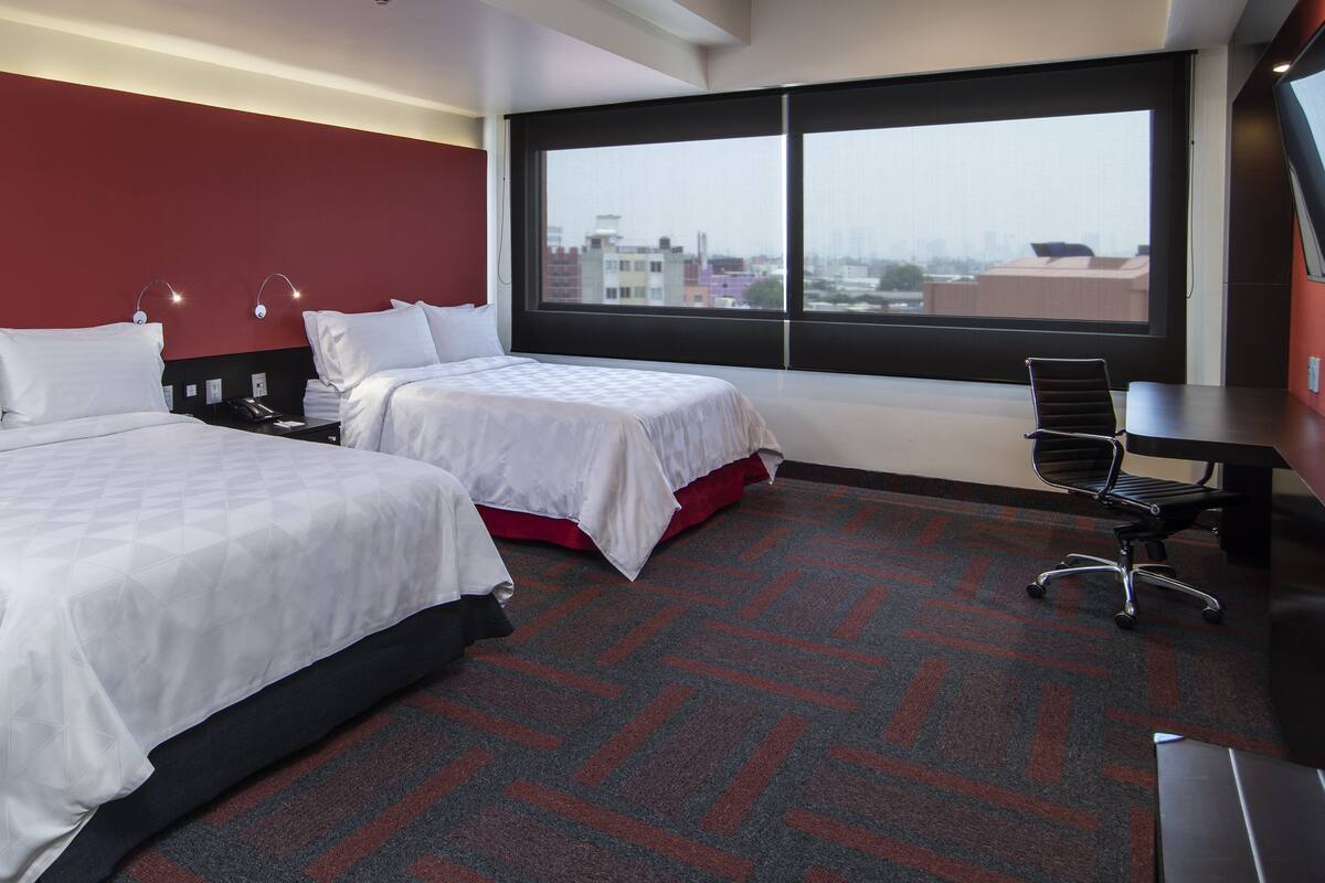 A hotel room with two beds, a desk, and a large window offering a city view.
