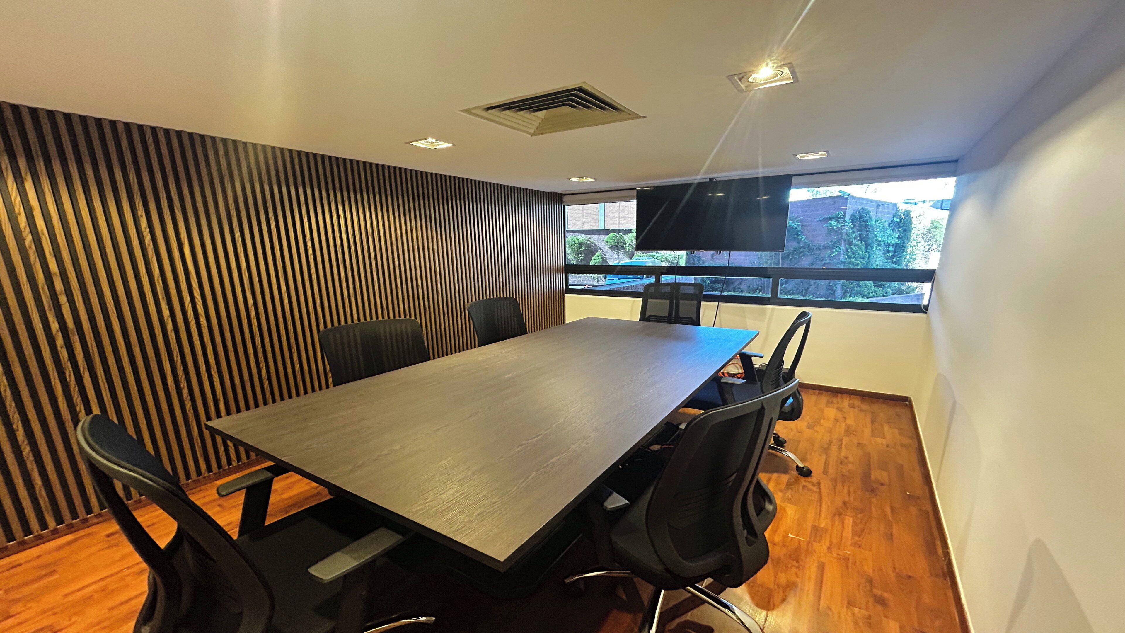 A conference room with a long table, black chairs, a TV, and a window with a view of trees.