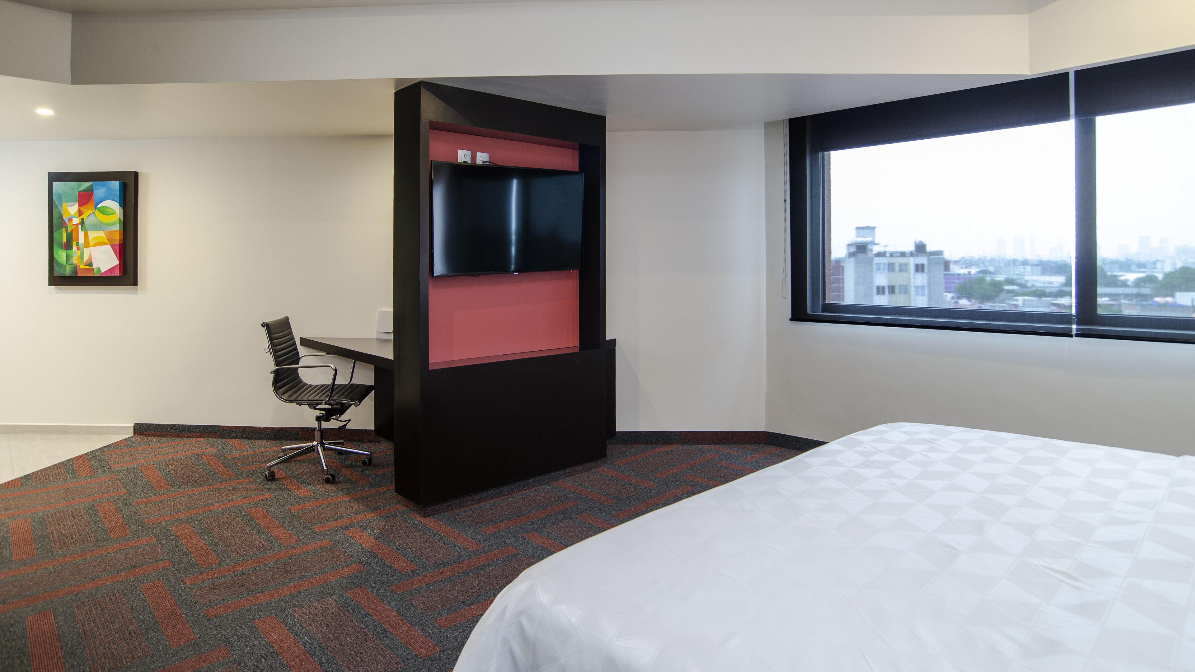 Habitación de hotel con cama, escritorio con silla, un mueble rojo de almacenamiento, ventana con vista a la ciudad y un cuadro de colores en la pared.