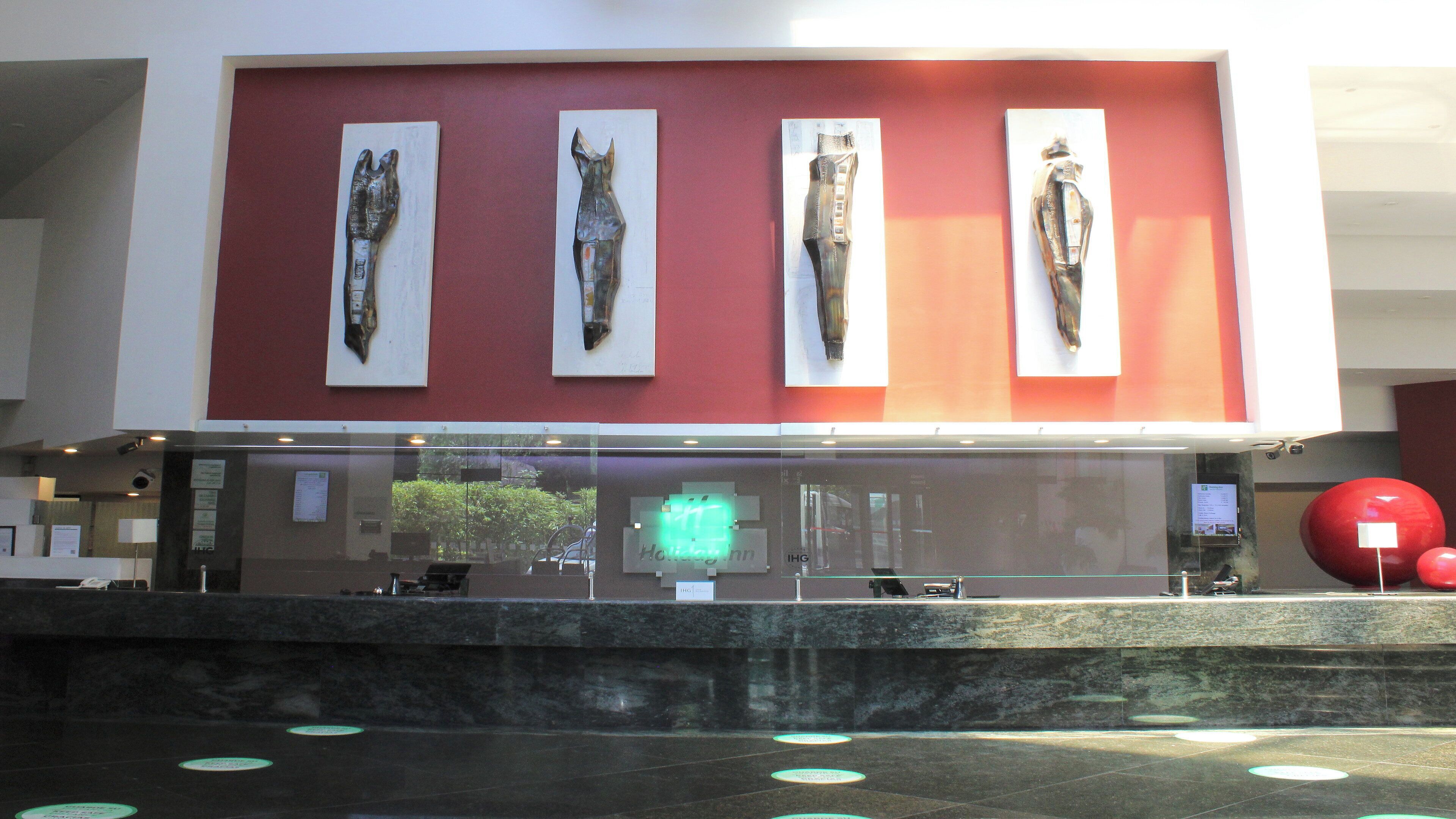 A reception area with a modern design, featuring a red wall and a glass partition. There are four framed artworks displayed above the reception desk.