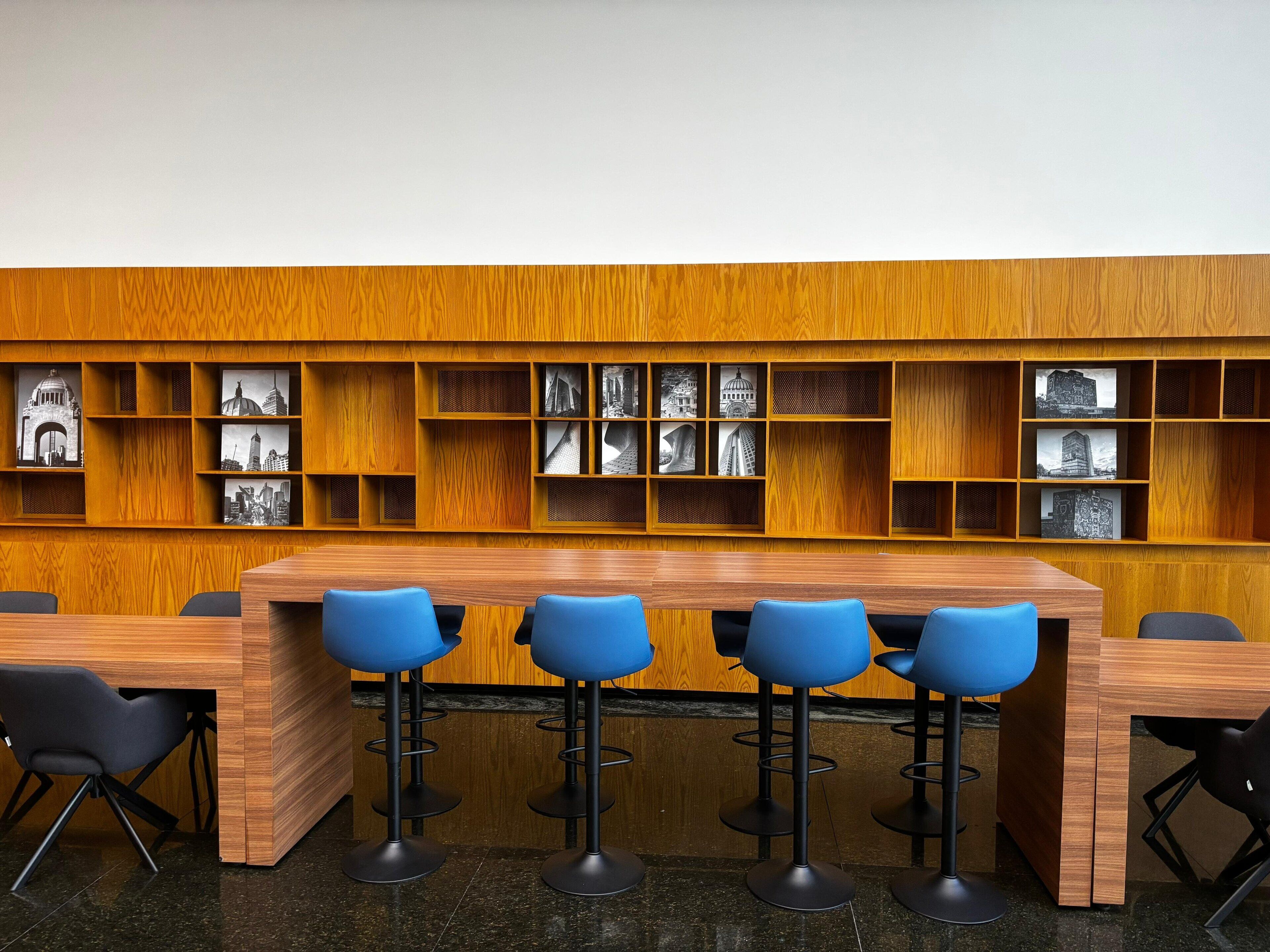 A modern library with a wooden bookshelf containing black and white photographs, a long wooden table with four blue bar stools, and black chairs.