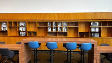 A modern library with a wooden bookshelf containing black and white photographs, a long wooden table with four blue bar stools, and black chairs.