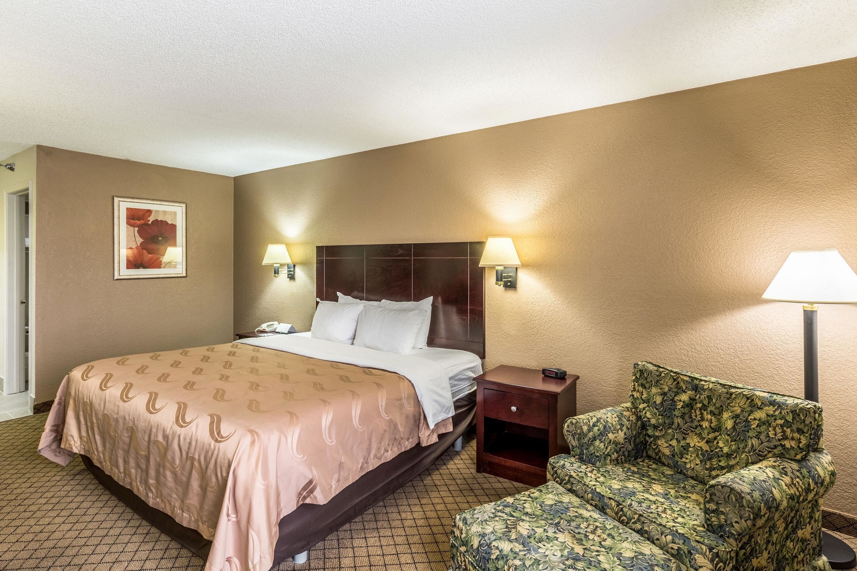 Una habitación de hotel con cama, lámparas de noche, mesita de noche, un sillón estampado con motivos florales y un cuadro en la pared.