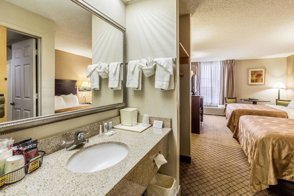 A hotel bathroom with a sink, mirror, and towel rack. There are two beds in the adjacent room.