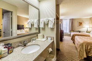 A hotel bathroom with a sink, mirror, and towel rack. There are two beds in the adjacent room.