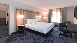 A hotel room with a large bed, bedside tables, a desk, and a chair. There is a window with curtains and a wall-mounted lamp.