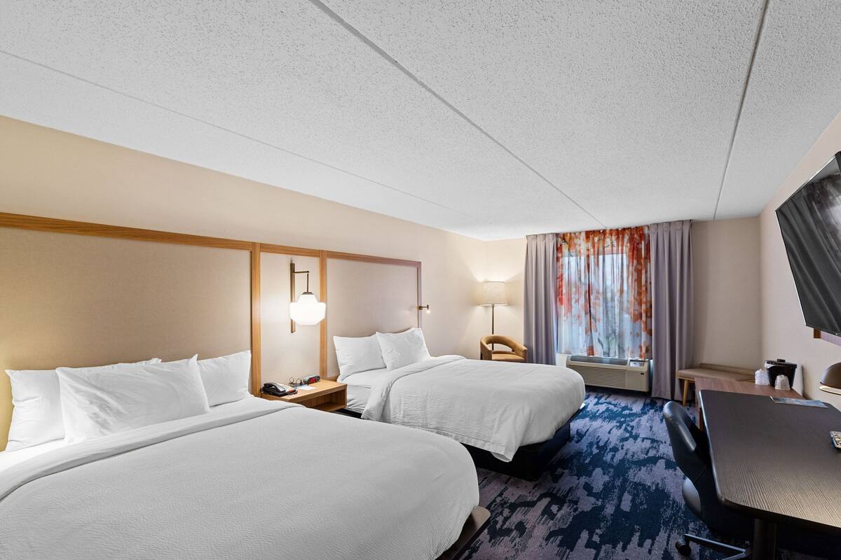Ett hotellrum med två sängar, ett skrivbord, en stol, en tv och ett fönster med gardiner.