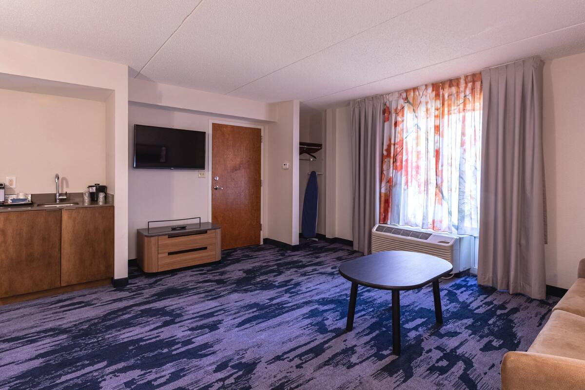 Ett hotellrum med ett litet kök, en platt-TV, ett runt bord, en soffa och ett stort fönster med gardiner.