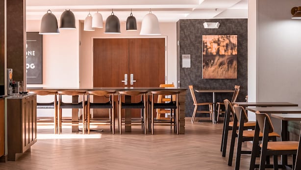 A modern restaurant with wooden tables and chairs, pendant lighting, and a large wooden door in the background.