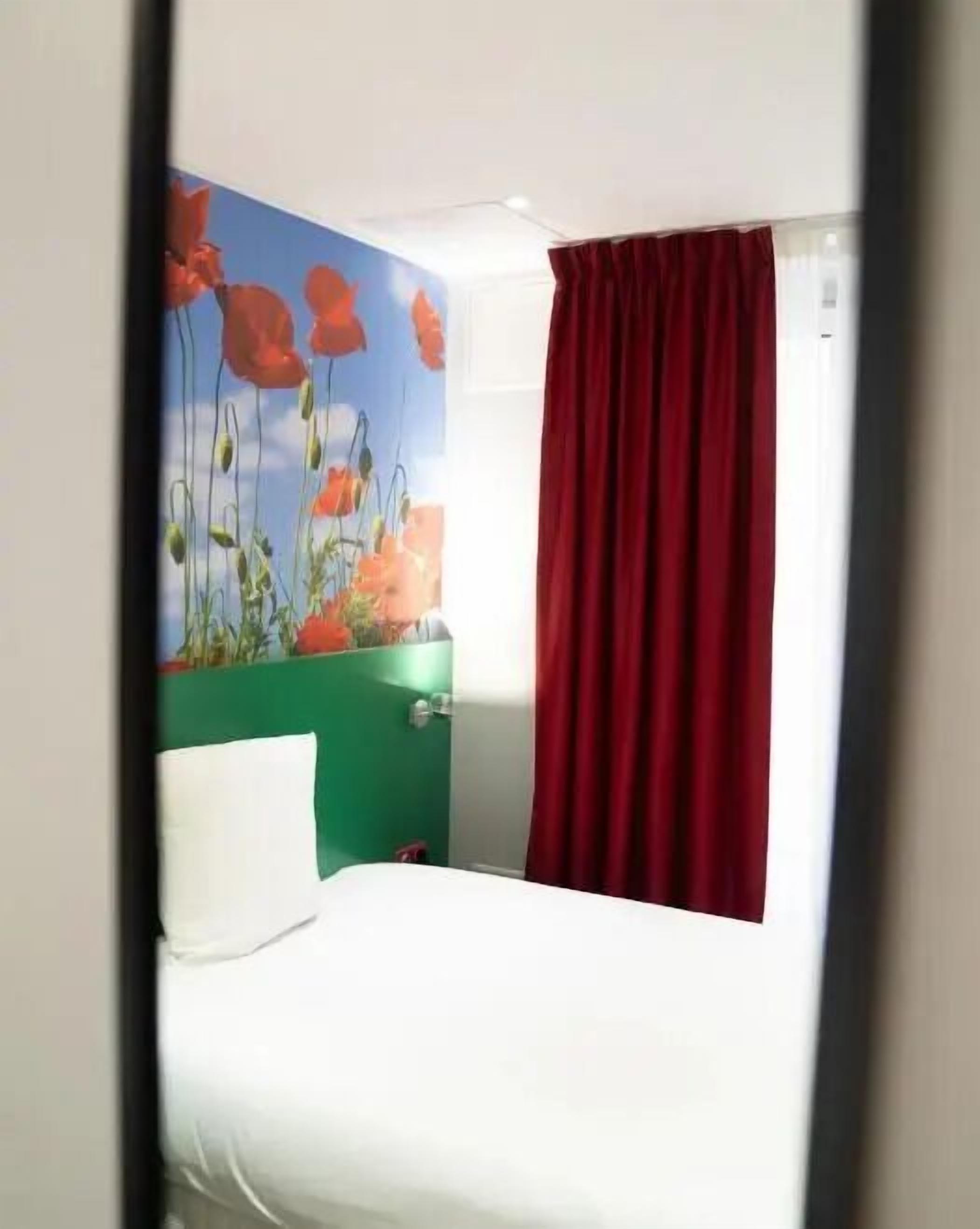 A hotel room with a bed, a green wall, a red curtain, and a painting of flowers.