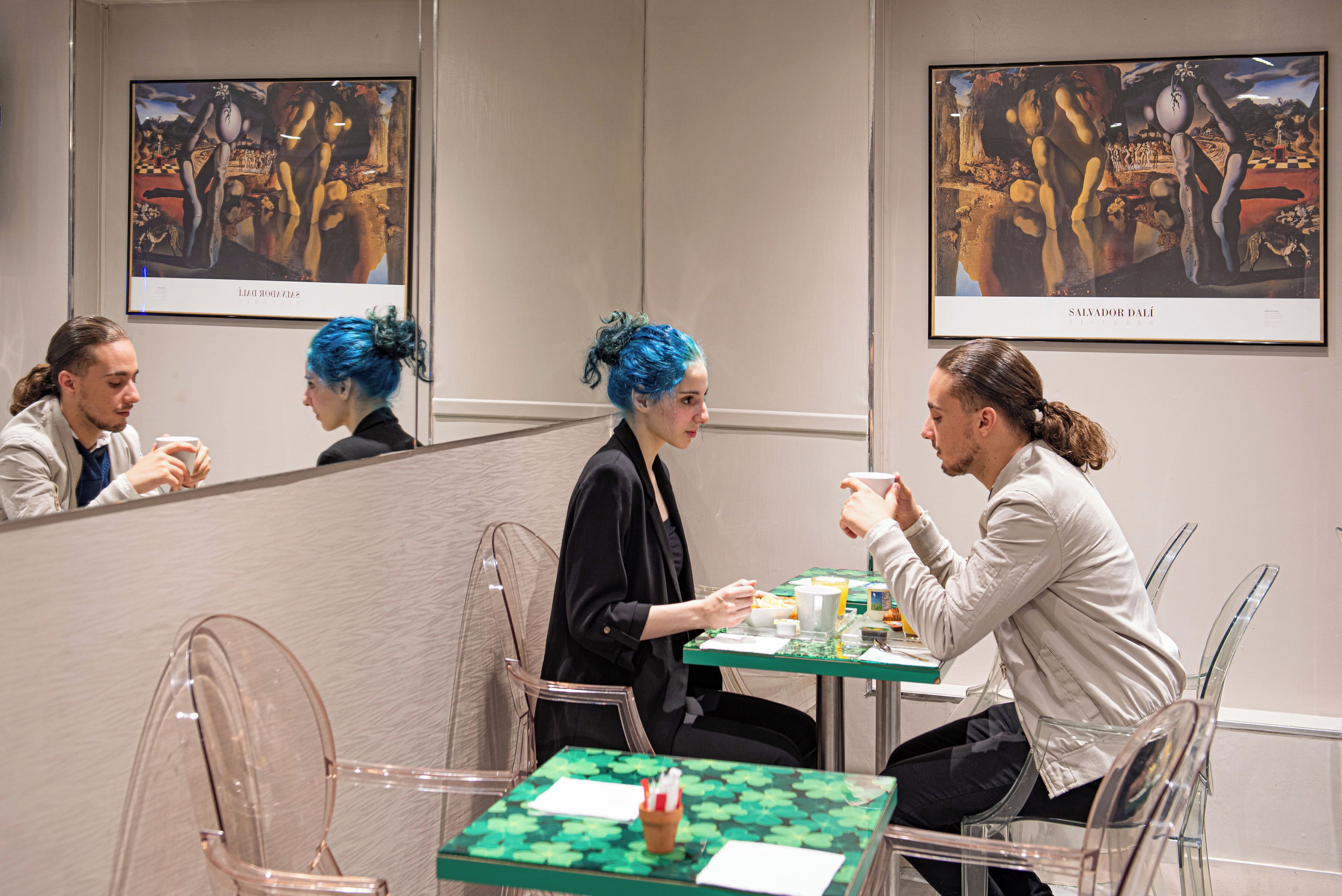 A dining area with clear glass chairs and tables, framed artwork on the walls, and a green tablecloth.